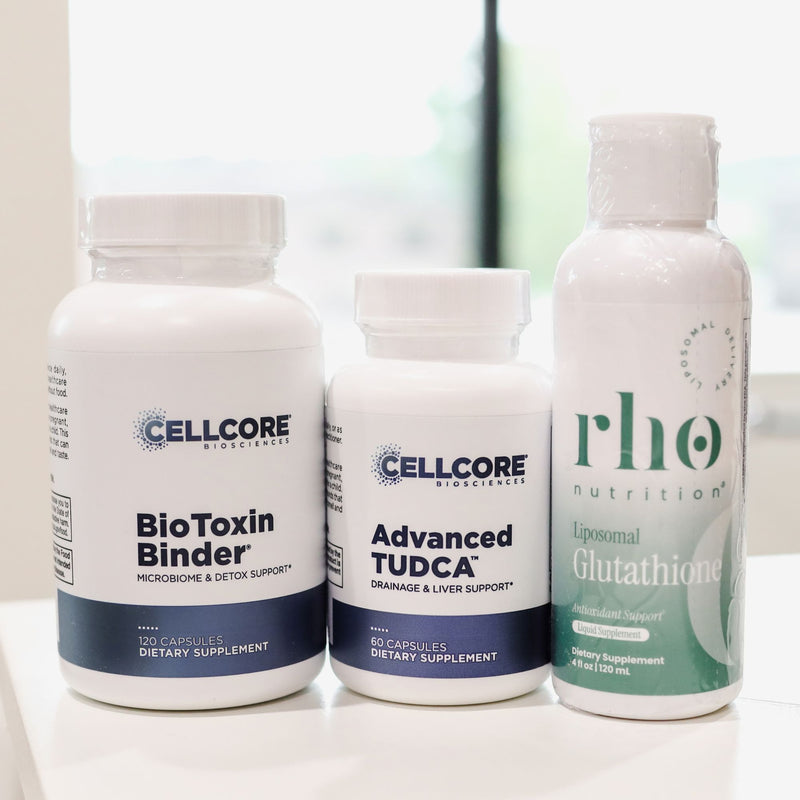 Three dietary supplement containers on a white surface: a tall white bottle labeled Cellcore BioToxin Binder, a shorter white bottle labeled Cellcore Advanced TUDCA, and a white bottle with a green label from Rhone Nutrition labeled Liposomal Glutathione.
