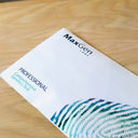 A white MaxGen Labs Professional Comprehensive Genetics Test booklet lies diagonally on a light wood grain surface. The booklet features blue and teal text and a stylized fingerprint graphic in the bottom right corner.