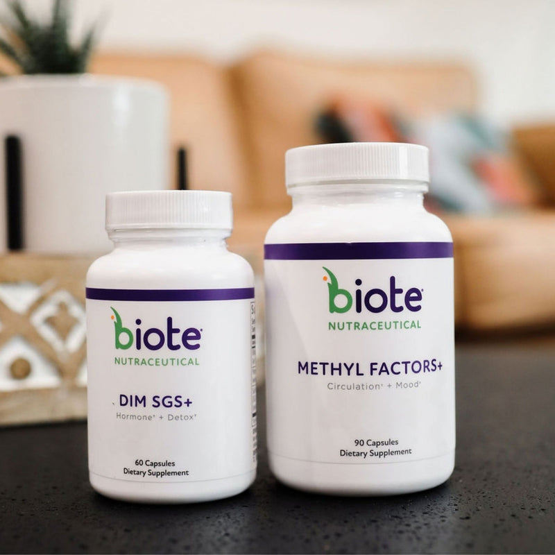 Two white biote Nutracetical dietary supplement bottles on a dark tabletop with a blurred living room background. The smaller bottle is labeled DIM SGS+ and the larger bottle is labeled Methyl Factors+.