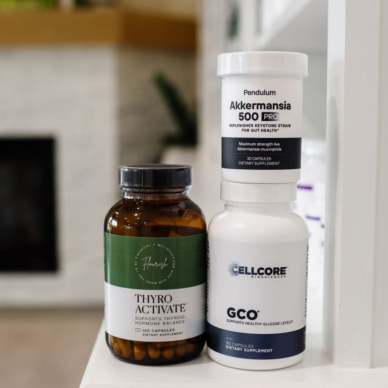Three dietary supplement bottles standing on a white surface in a softly blurred indoor setting: an amber bottle labeled Thyro Activate, a white bottle labeled Pendulum Akkermansia 500 EC, and a white bottle labeled Cellcore GCO.