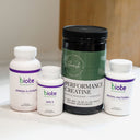 Four dietary supplement containers from Biote Nutritional arranged on a white surface: a large black tub labeled Performance Creatine, a white bottle labeled Omega 3+ COQ10, a smaller white bottle labeled ADK 5, and a white bottle labeled Methyl Factors+. The background shows a softly blurred indoor setting with warm wood tones.