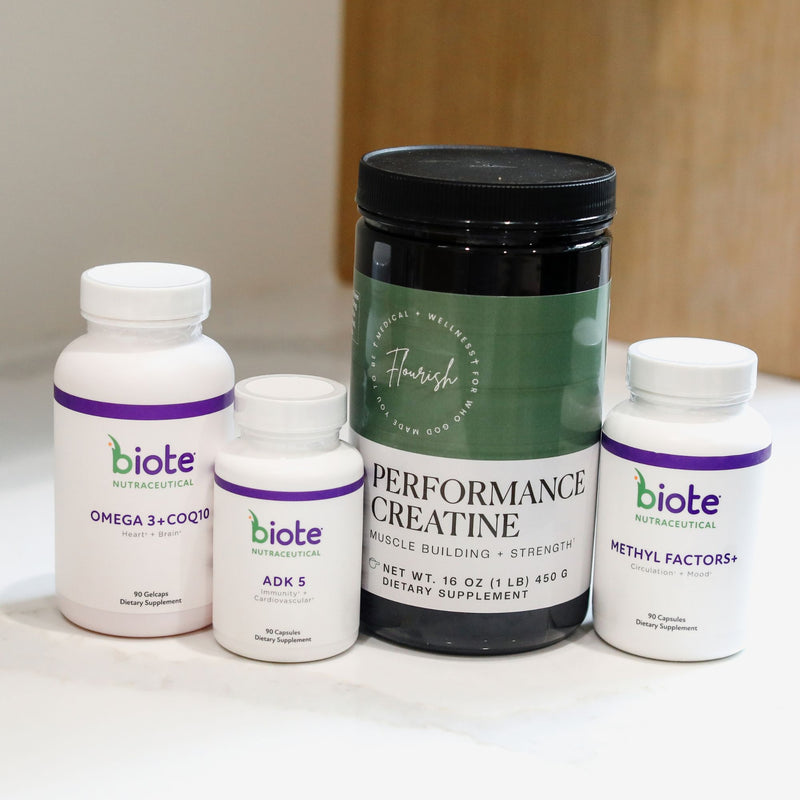 Four dietary supplement containers from Biote Nutritional arranged on a white surface: a large black tub labeled Performance Creatine, a white bottle labeled Omega 3+ COQ10, a smaller white bottle labeled ADK 5, and a white bottle labeled Methyl Factors+. The background shows a softly blurred indoor setting with warm wood tones.