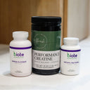 Three dietary supplement bottles grouped together on a light countertop with a blurred wood background. The center bottle is a large black tub labeled Performance Creatine, flanked by two white bottles labeled biote Nutracutical Omega 3+ COQ10 and biote Nutracutational Methyl Factor+.