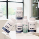 Five white supplement bottles from CellCore Nutrition arranged on a white surface in front of a softly blurred window background. The products include BioToxin Binder, CT-Minerals, Advanced TUDDCA, RhO Enzymed Glutathione, and BC-ATP.