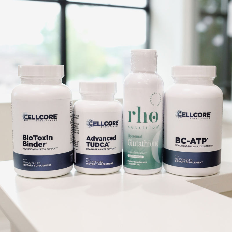Four white supplement bottles from Cellcore Nutrition arranged on a white surface in front of a blurred window background. The bottles include BioToxin Binder, Advanced TUDCA, L-Theanine, and BC-ATP.