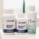 Three dietary supplement containers on a white surface: a tall white bottle labeled Cellcore BioToxin Binder, a shorter white bottle labeled Cellcore Advanced TUDCA, and a white bottle with a green label from Rhone Nutrition labeled Liposomal Glutathione.
