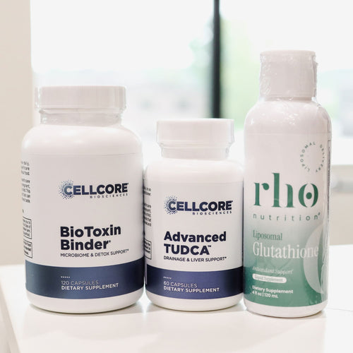 Three dietary supplement containers on a white surface: a tall white bottle labeled Cellcore BioToxin Binder, a shorter white bottle labeled Cellcore Advanced TUDCA, and a white bottle with a green label from Rhone Nutrition labeled Liposomal Glutathione.