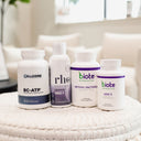 Four dietary supplement bottles arranged on a white woven stool in a bright, modern living room setting. The products include Cellcore BC-ATP, Rheus NAD+ (a liquid bottle), Biote Methyl Factorals (a white bottle with purple accents), and Biote ADK 5.