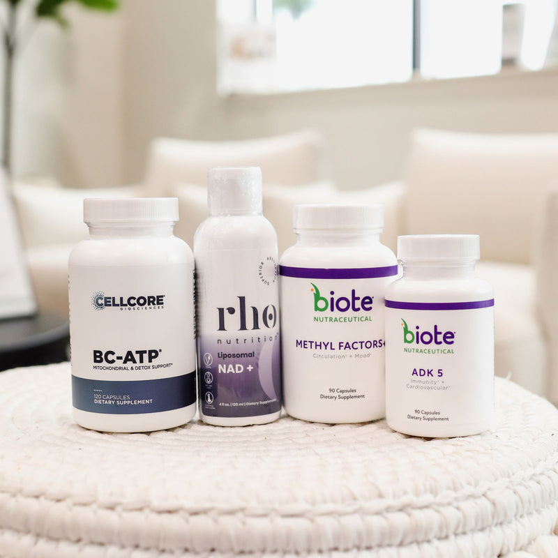 Four dietary supplement bottles arranged on a white woven stool in a bright, modern living room setting. The products include Cellcore BC-ATP, Rheus NAD+ (a liquid bottle), Biote Methyl Factorals (a white bottle with purple accents), and Biote ADK 5.