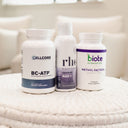 Three white dietary supplement bottles standing on a textured cream blanket in a softly blurred indoor setting. The bottles are labeled Cellcore BC-ATP, Rhe Liposomal NAD+ by Rhe Nutrition, and Biote Methyl Factors+.