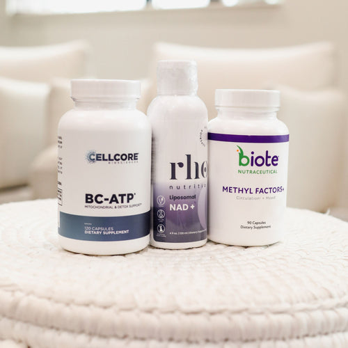 Three white dietary supplement bottles standing on a textured cream blanket in a softly blurred indoor setting. The bottles are labeled Cellcore BC-ATP, Rhe Liposomal NAD+ by Rhe Nutrition, and Biote Methyl Factors+.