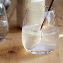 A clear stemless wine glass filled with a pale frothy liquid, set on a light wooden table with a metal spoon resting inside and a blurred glass in the background.