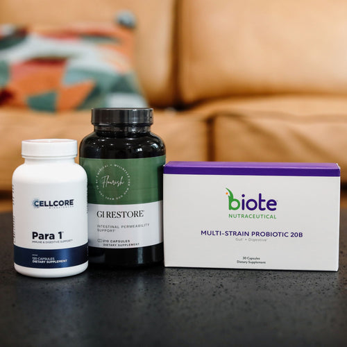 Three dietary supplement products arranged on a dark countertop in front of a blurred living room background: a white CELLCORE Para 1 bottle, a dark green GI Restore bottle, and a white biote Nutracetical Multi-Strain Probiotic 208 box.