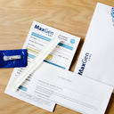 MaxGen Labs genetic test kit components arranged on a light wood surface, including printed forms, a white plastic pipette, a blue vial packet, and branded envelopes.
