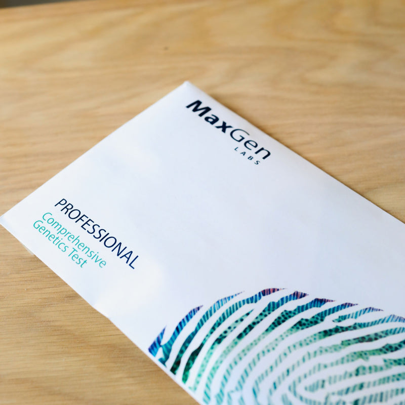 A white MaxGen Labs Professional Comprehensive Genetics Test booklet lies diagonally on a light wood grain surface. The booklet features blue and teal text and a stylized fingerprint graphic in the bottom right corner.