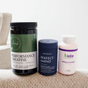 Three supplement containers arranged on a textured beige surface against a soft light gray background: a black jar labeled Flavour Performance Creatine, a navy blue canister labeled Bodyhealth Perfect Amino, and a pale pink bottle labeled biote Omega 3 + CoQ10.
