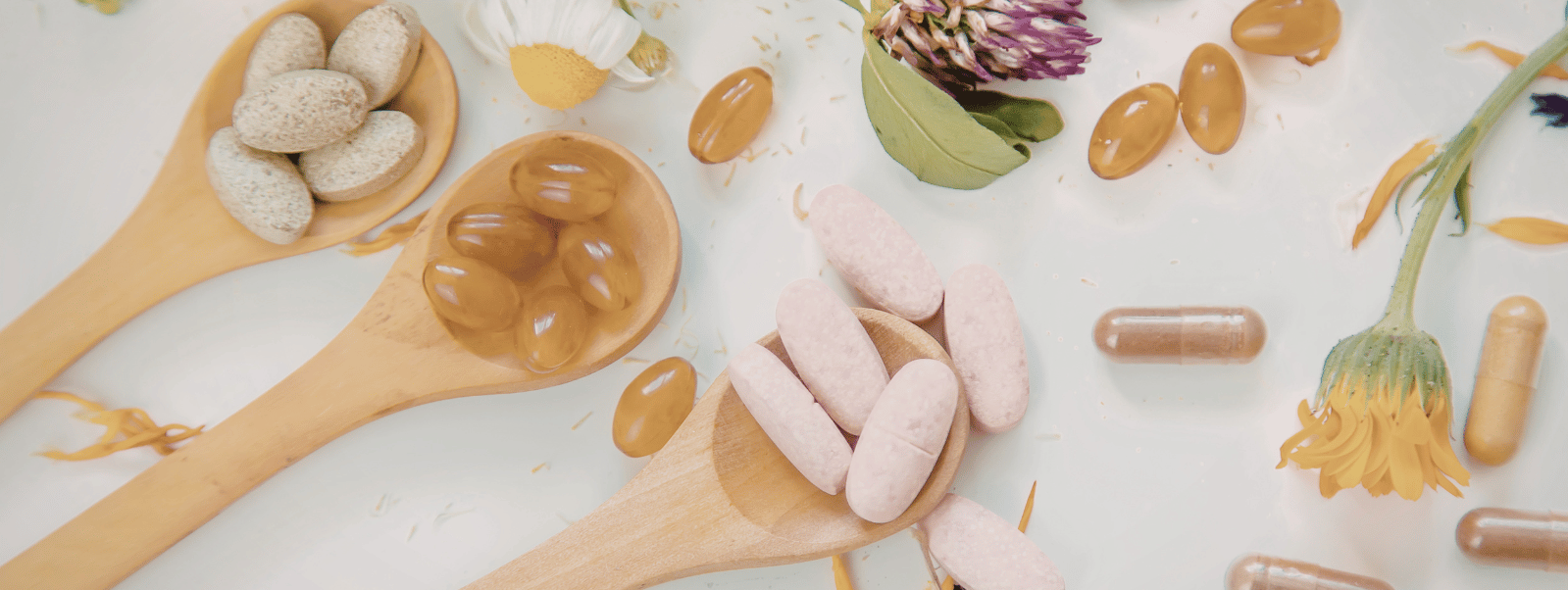 Spoons filled with various pills and capsules on a light background with flowers and herbs.