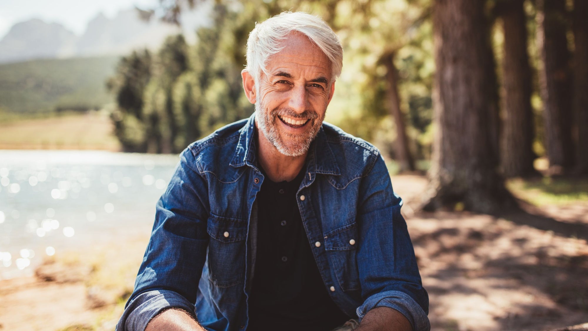 gray-haired man smiling outdoors sitting on a rock near a lake