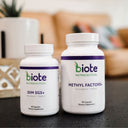 Two white biote Nutracetical dietary supplement bottles on a dark tabletop with a blurred living room background. The smaller bottle is labeled DIM SGS+ and the larger bottle is labeled Methyl Factors+.