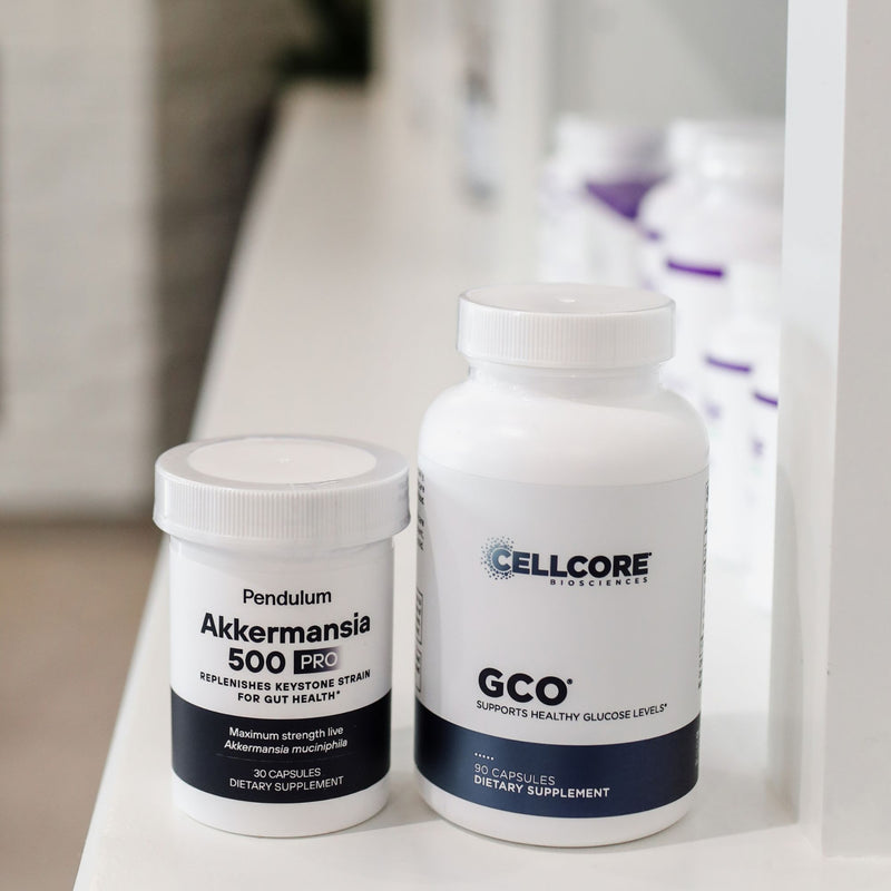 Two white supplement bottles on a white shelf: a smaller Pendulum Akkermansia 500 Pro bottle and a larger Cellcore GCO bottle, with a softly blurred background of more bottles.